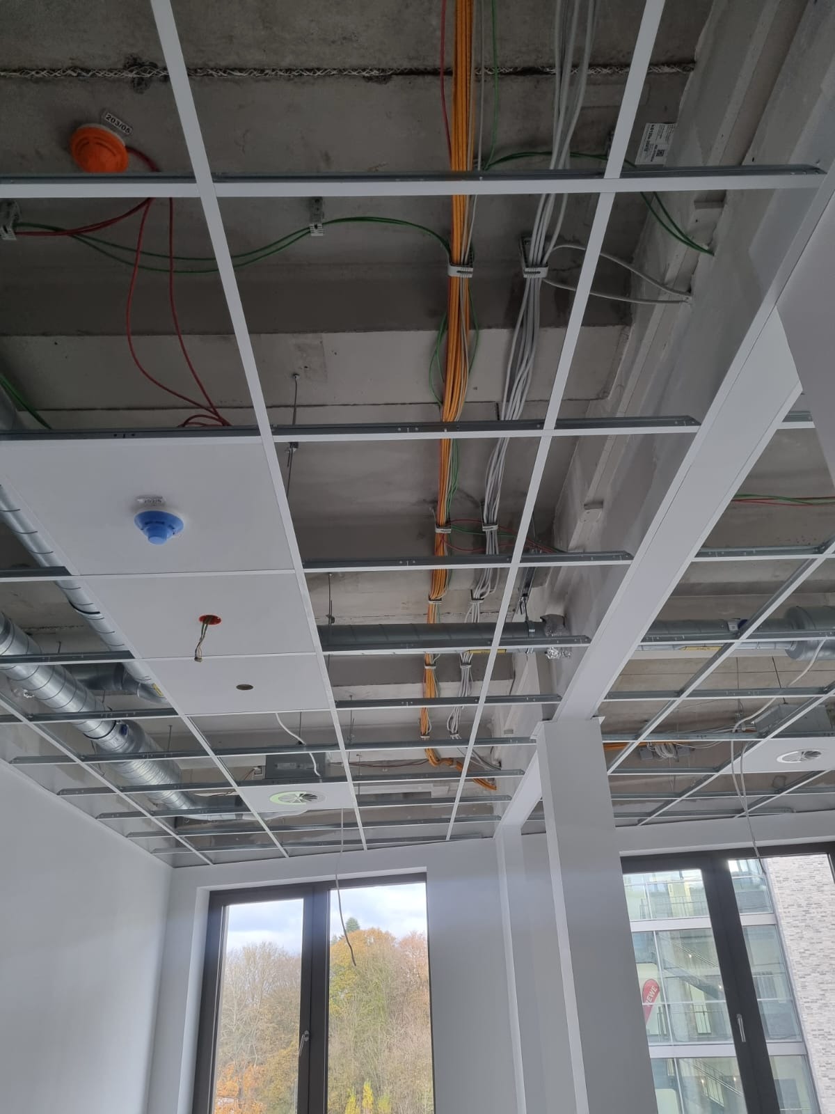 Sufit podwieszany w trakcie montażu z widocznymi instalacjami elektrycznymi i wentylacyjnymi. Widok z dołu na metalową konstrukcję i okablowanie.