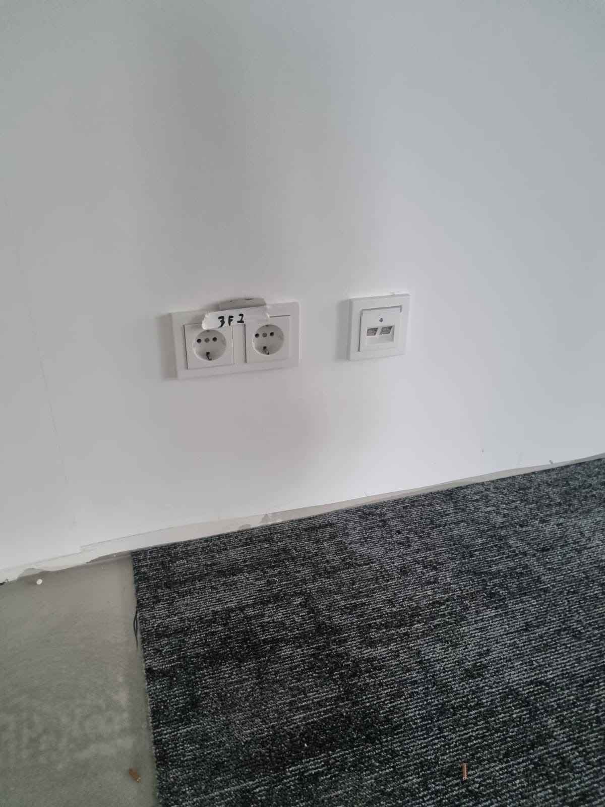 Biała ściana z dwoma gniazdkami elektrycznymi i gniazdem sieciowym. Na ścianie widoczny napis '3F2'. Na podłodze szara wykładzina dywanowa.