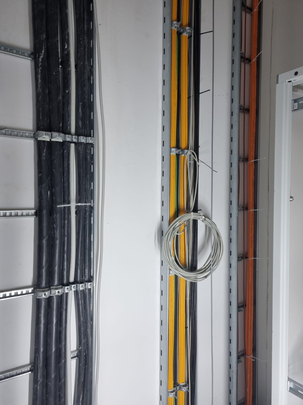 Różnokolorowe przewody elektryczne, czarne, żółte i pomarańczowe, prowadzone w metalowych korytkach na białej ścianie. Część przewodów zwinięta w pętlę.