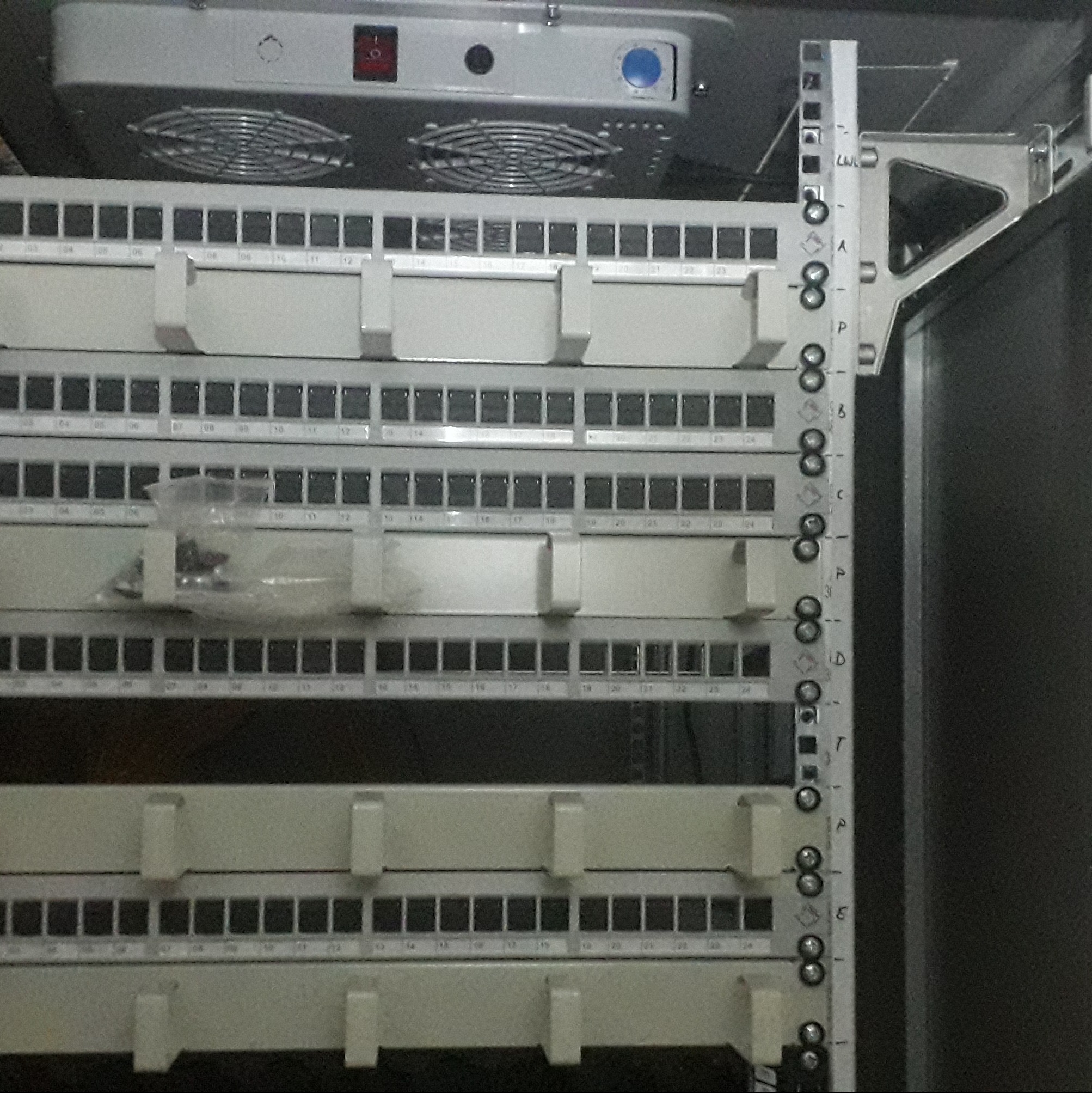 Panel krosowniczy w szafie rack z numerowanymi portami i wentylatorem chłodzącym. Widoczne kable i elementy montażowe. Oświetlenie górne, kąt ujęcia od dołu.