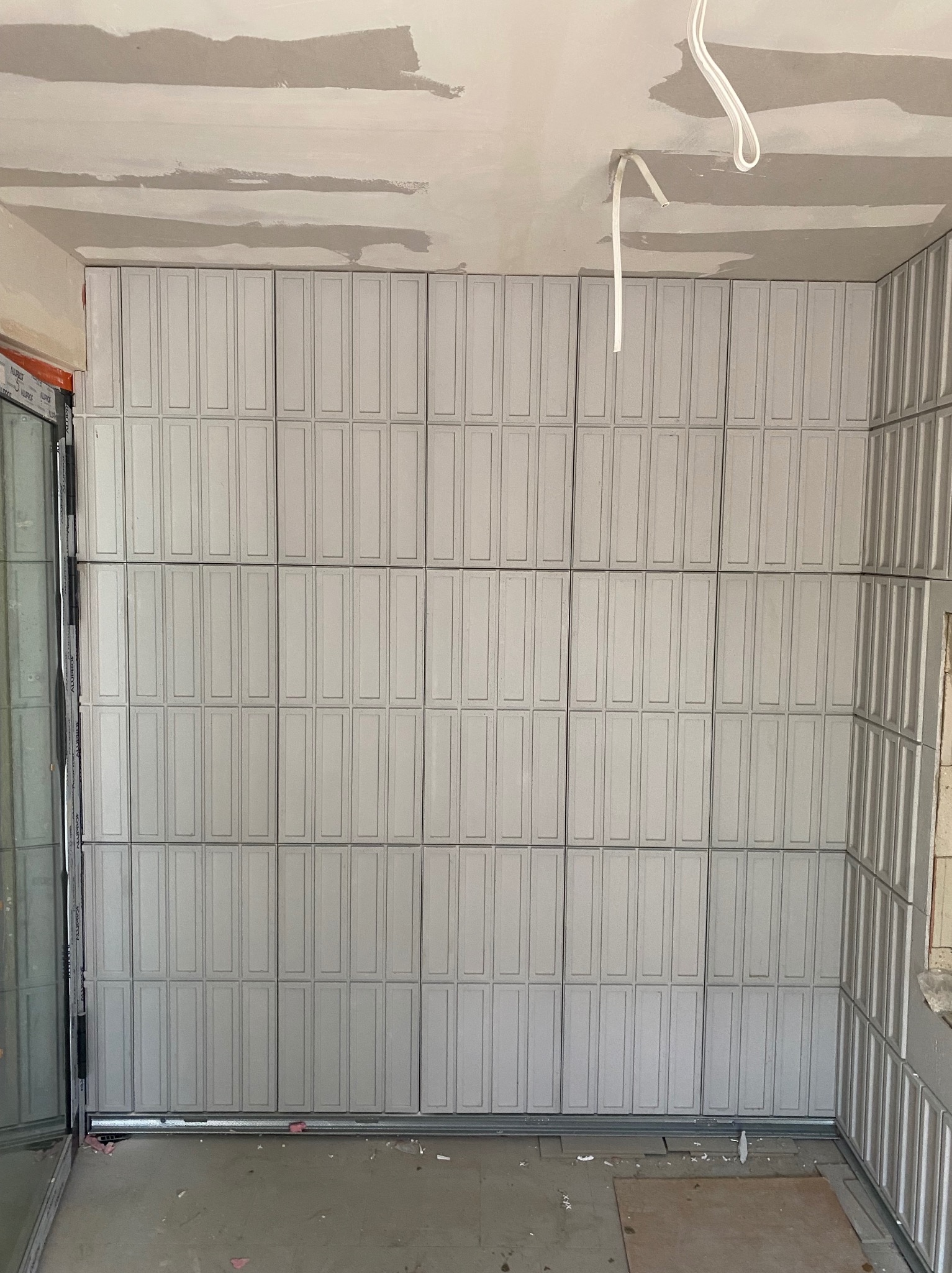 Ściana w trakcie wykańczania z szarymi, prostokątnymi płytkami imitującymi panele. Widoczne niedoskonałości na suficie i wystające kable. Prace wykończeniowe w pomieszczeniu.