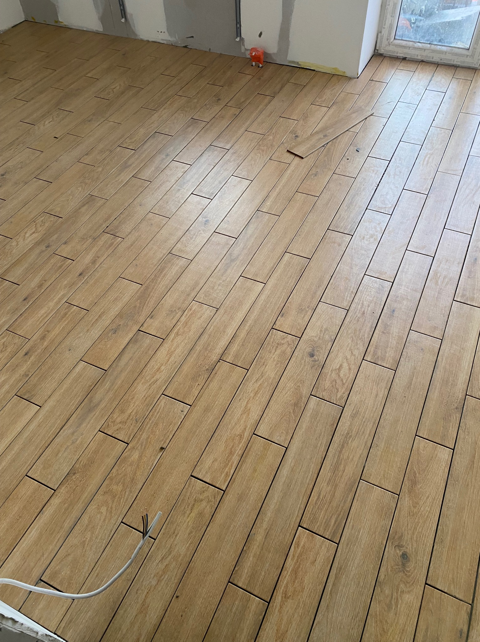 Ułożona podłoga z płytek imitujących drewno w odcieniach beżu i brązu. Widoczny fragment ściany i okna. Jedna płytka leży luzem. W rogu wystają przewody.