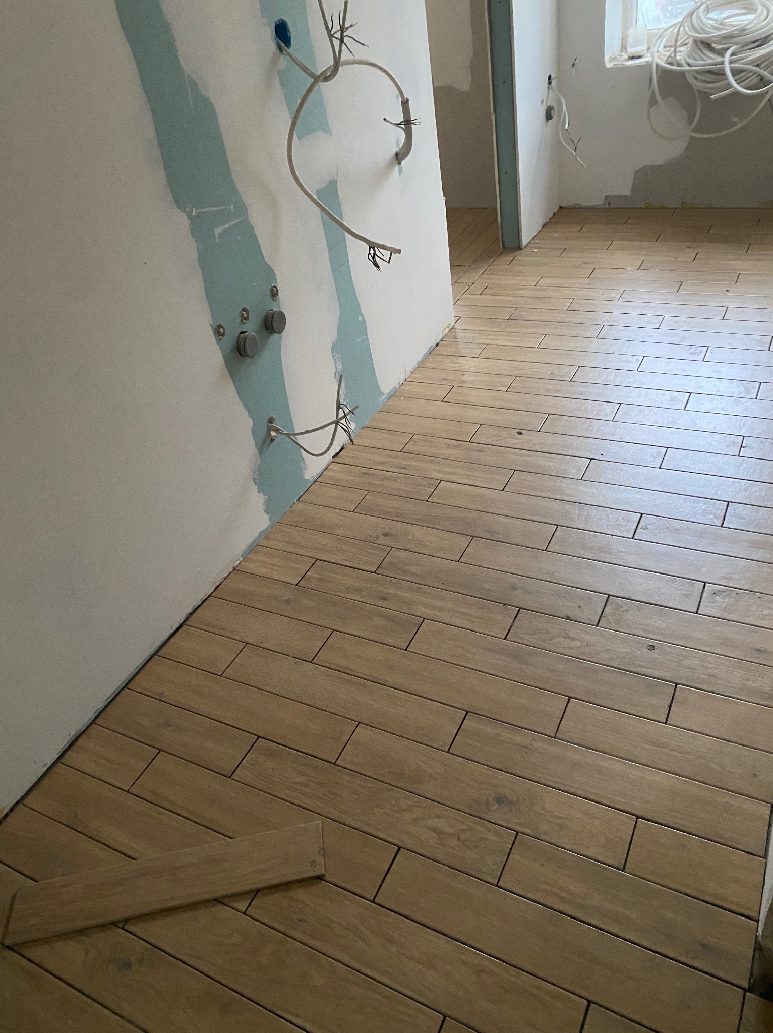 Ułożona podłoga z płytek imitujących drewno w łazience w trakcie remontu. Widoczne przygotowane ściany i wystające kable. Jedna płytka leży luzem na podłodze.