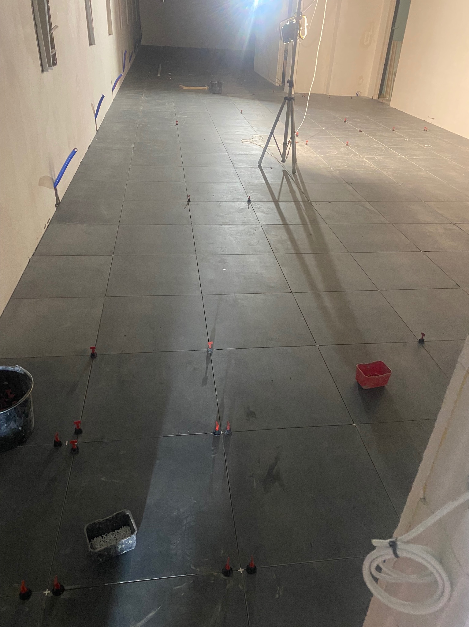 Układanie ciemnych, kwadratowych płytek podłogowych z użyciem poziomujących klipsów i klinów. Widoczny reflektor budowlany i fragment instalacji wodnej przy ścianie.