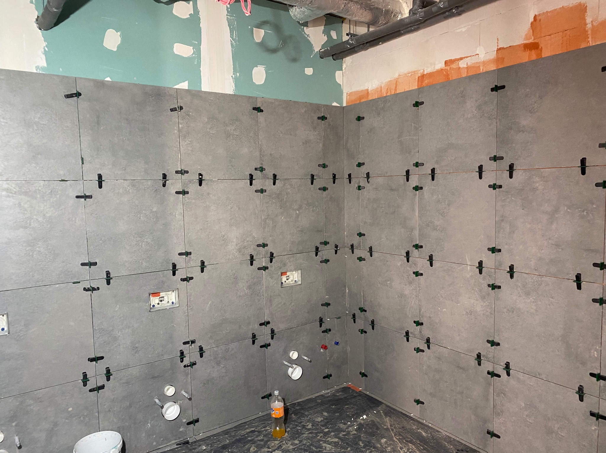 Szare płytki ścienne w łazience, świeżo ułożone z użyciem klipsów poziomujących. Widoczne instalacje wodne i elektryczne. Częściowo wykończone ściany.