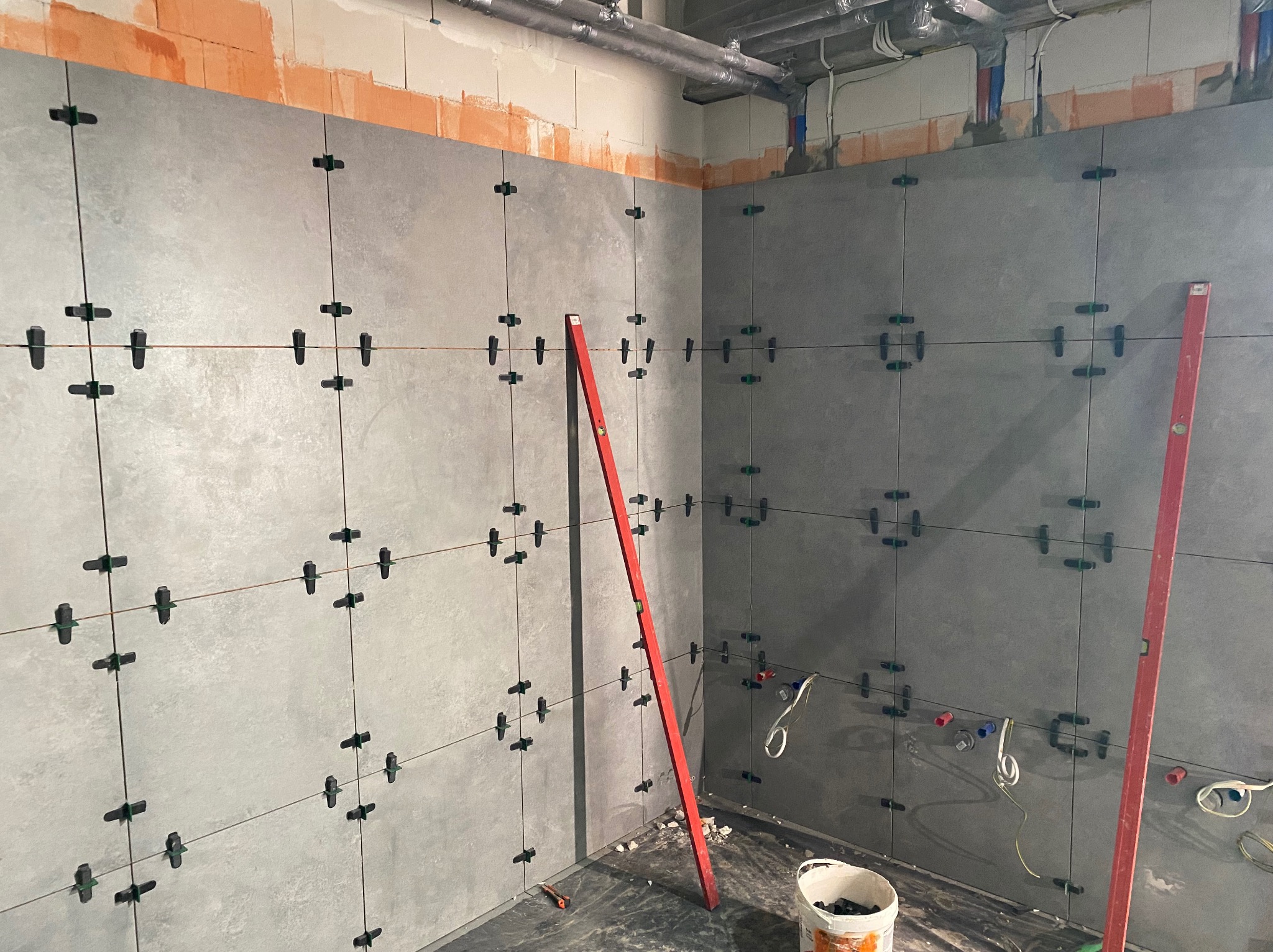 Szare płytki ścienne w trakcie układania, krzyżyki dystansowe, poziomica oparta o ścianę, widoczne instalacje i fragment podłogi z resztkami materiałów.