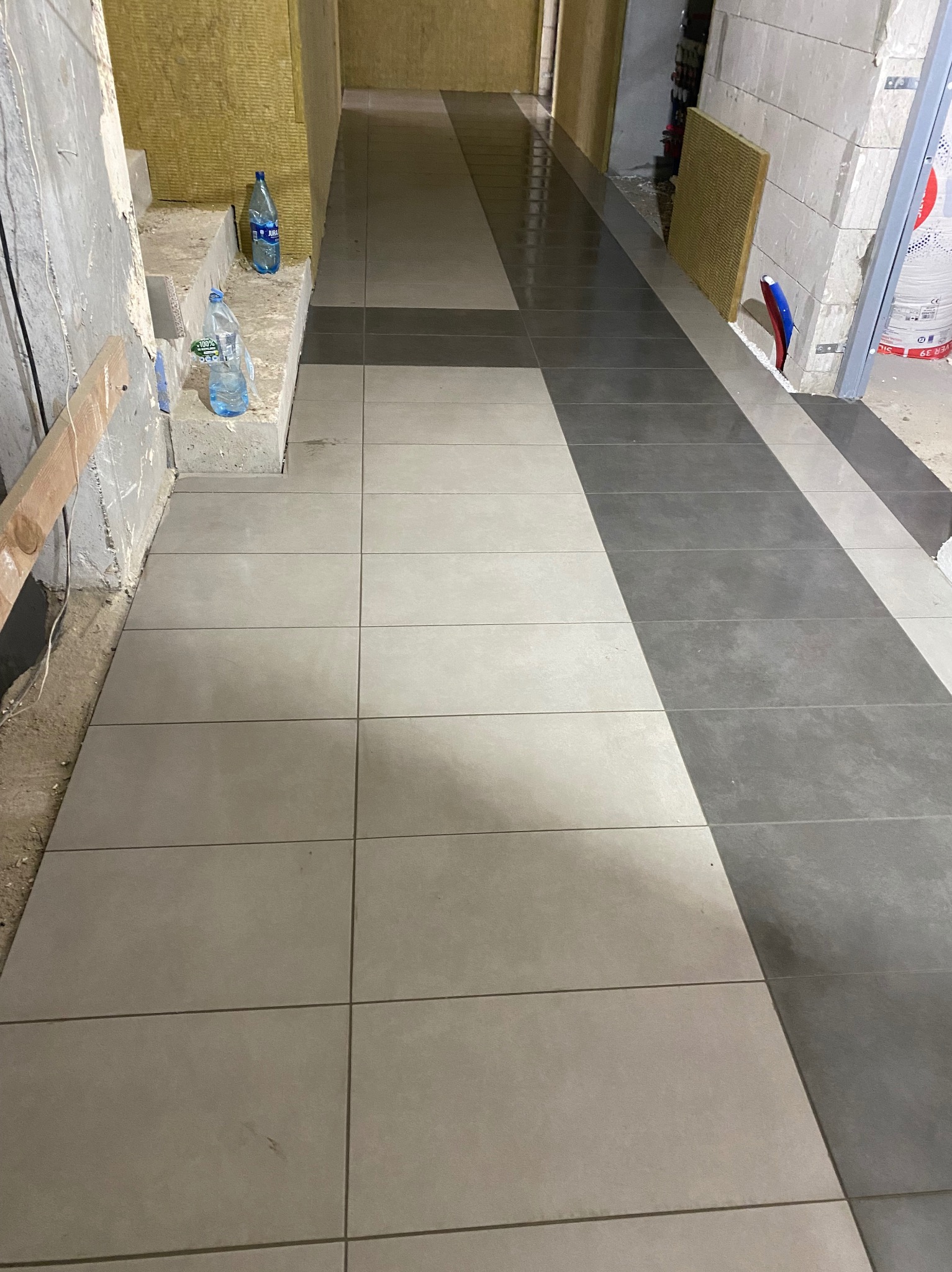 Ułożona podłoga z płytek w odcieniach szarości, z widocznymi fragmentami ścian w trakcie prac wykończeniowych i butelkami wody mineralnej w tle.