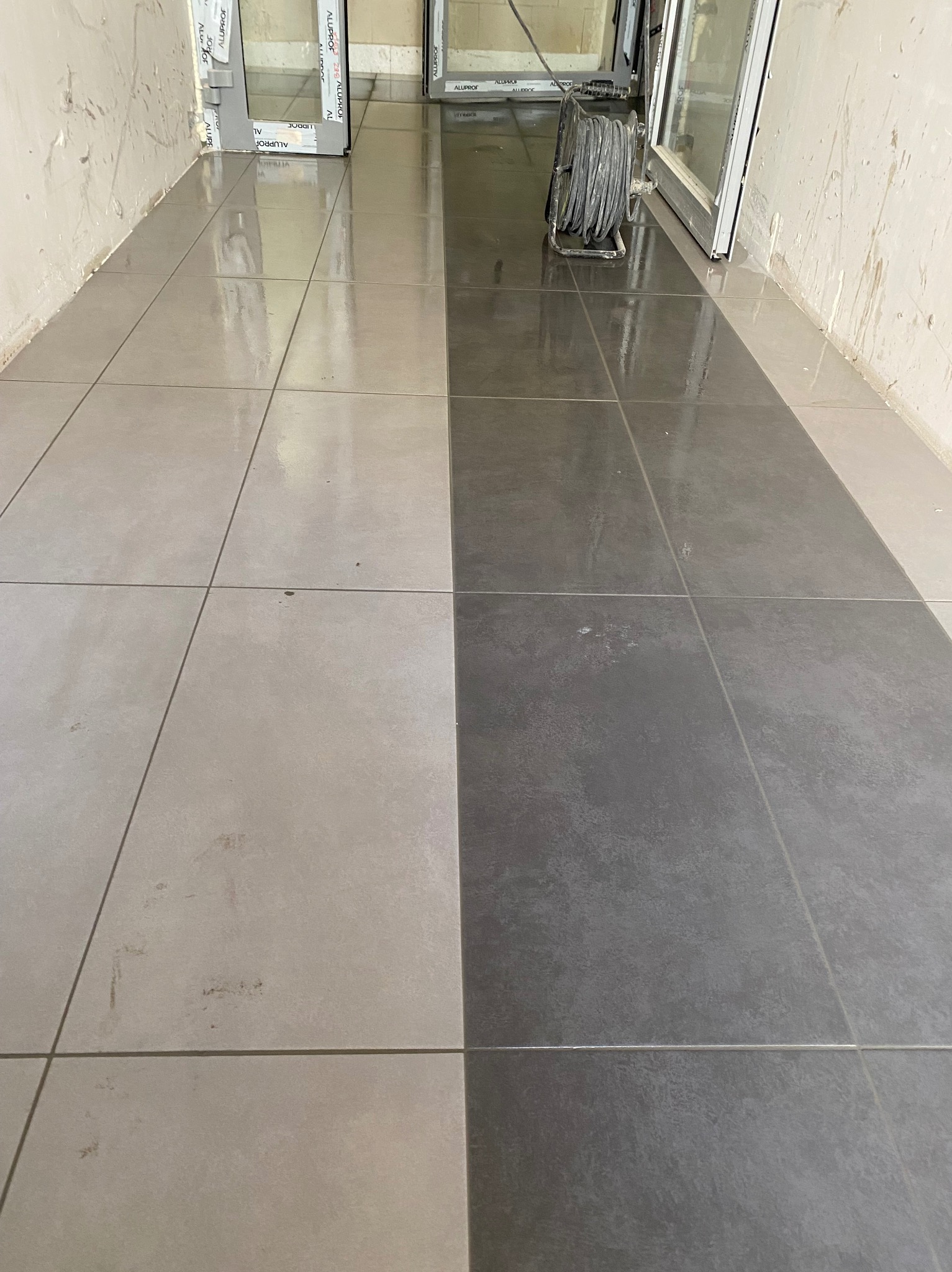 Ułożona podłoga z płytek w dwóch odcieniach szarości, z widocznym kablem zwiniętym na bębnie przy ścianie i oknem w tle. Widok z perspektywy podłogi.