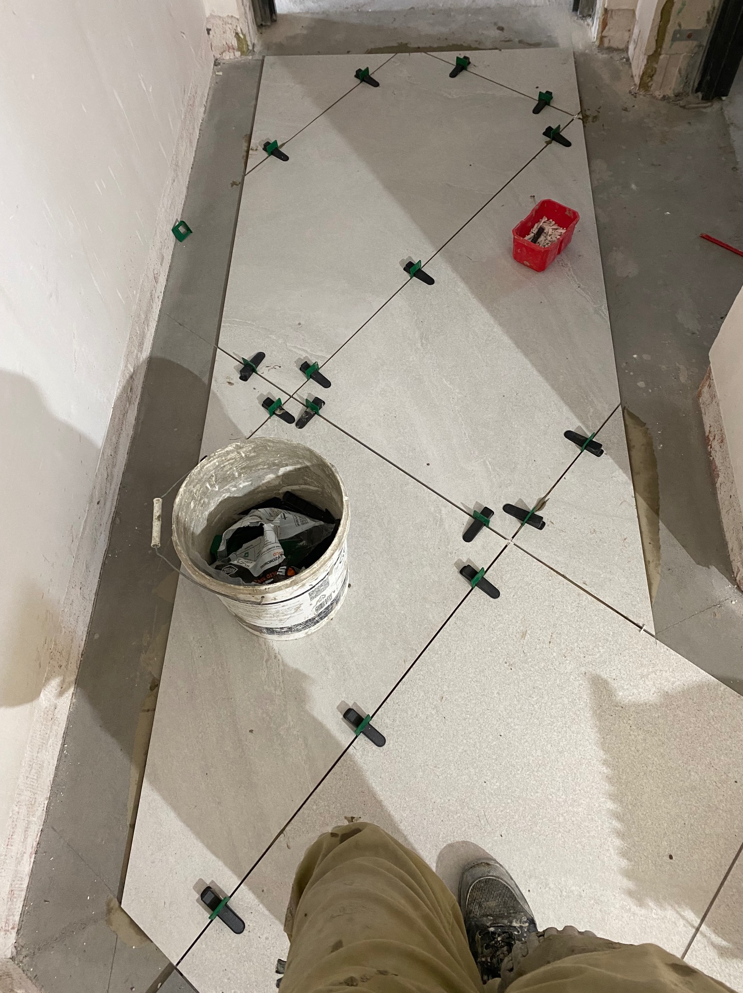 Układanie jasnych płytek podłogowych z użyciem systemu poziomowania. Widoczny fragment buta i wiadro z narzędziami. Prace w toku, surowy stan wykończenia ścian.