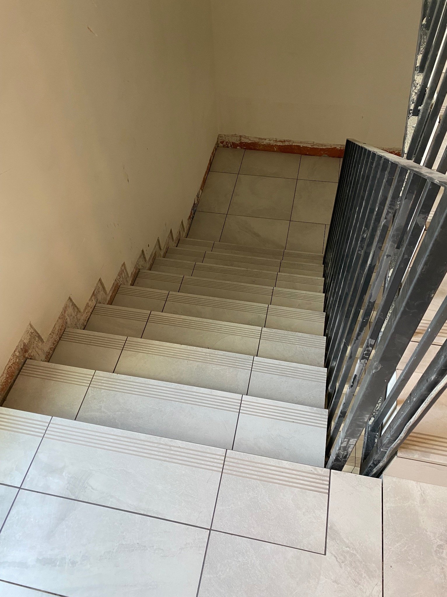 Wykończone schody z jasnymi, kwadratowymi płytkami z delikatnym wzorem, widok z góry. Fragment ściany z widocznymi śladami po wcześniejszych pracach. Metalowa barierka.