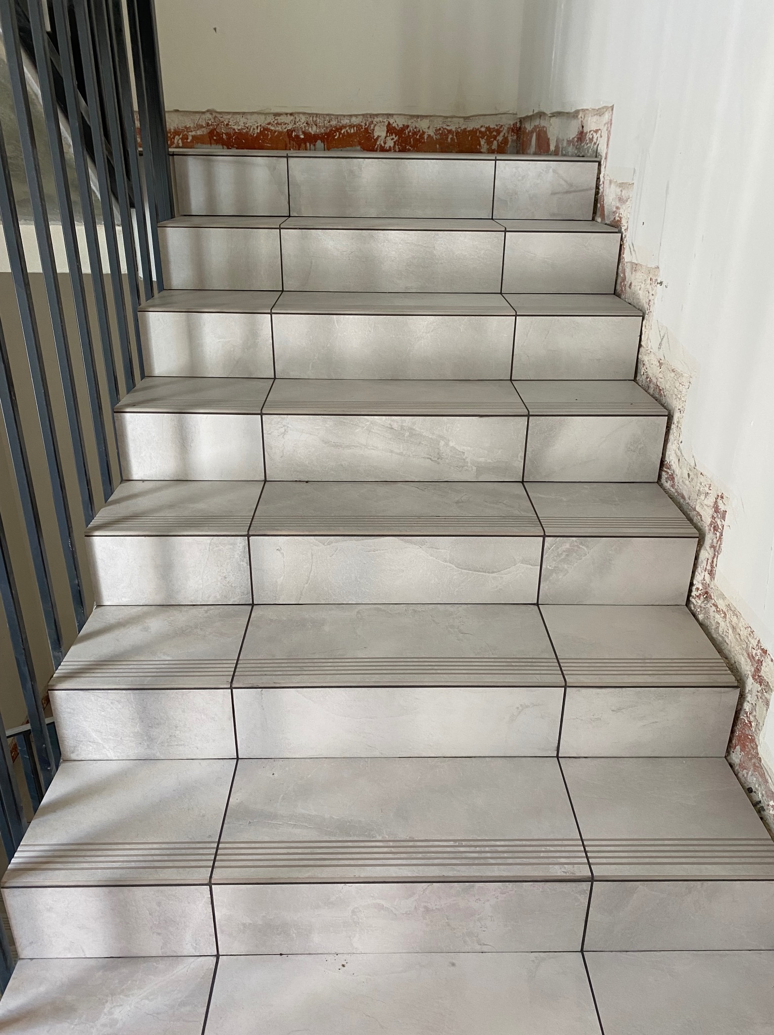 Wykończone schody z szarych płytek imitujących kamień, z ciemnymi fugami. Widoczne niedociągnięcia na ścianie przy schodach. Metalowa barierka po lewej stronie.