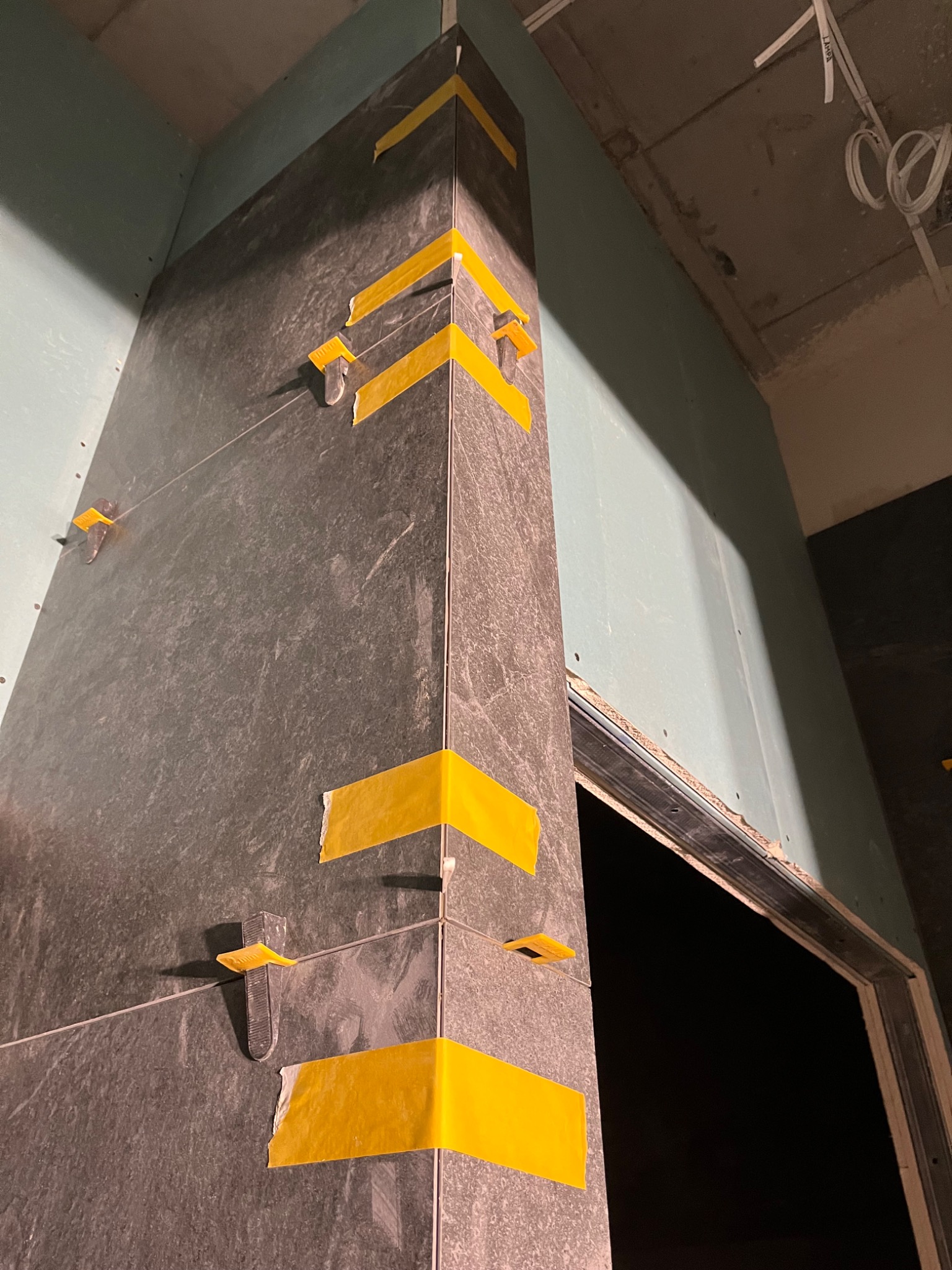 Układanie ciemnych płytek na ścianie z użyciem żółtej taśmy i klipsów poziomujących. Widoczny narożnik i fragment ściany z płyt gipsowo-kartonowych.