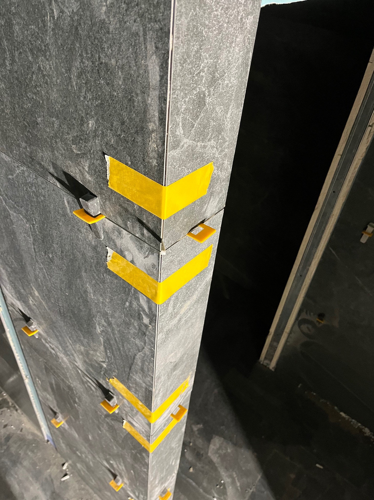 Montaż ciemnych, matowych płytek na ścianie z użyciem żółtej taśmy i klipsów poziomujących. Widoczny fragment ściany i podłogi w trakcie prac glazurniczych.