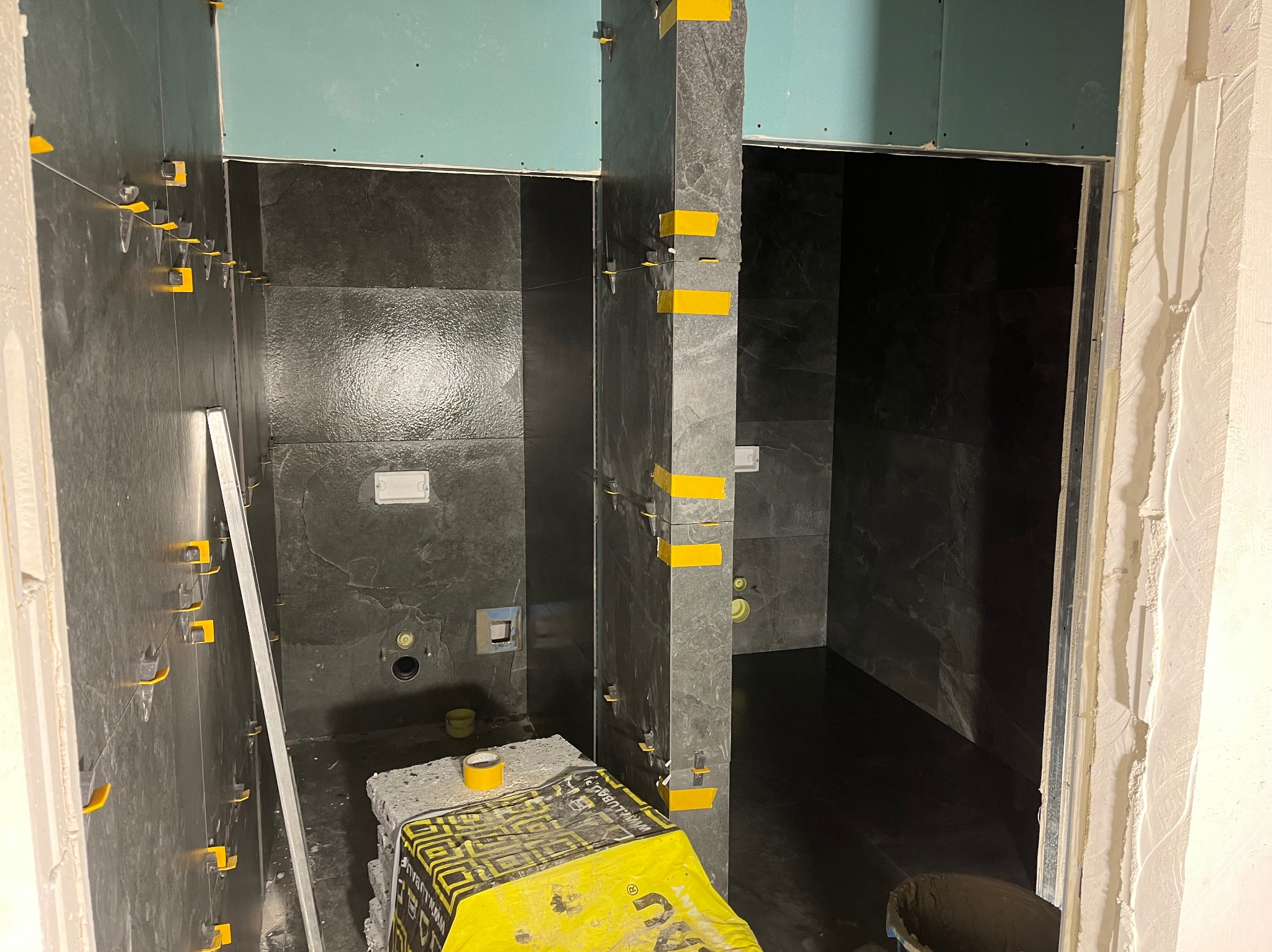 Układanie ciemnych, marmurkowych płytek w łazience w Ząbkach. Widoczne poziomice, paczka kleju i system poziomowania płytek. Etap prac wykończeniowych.