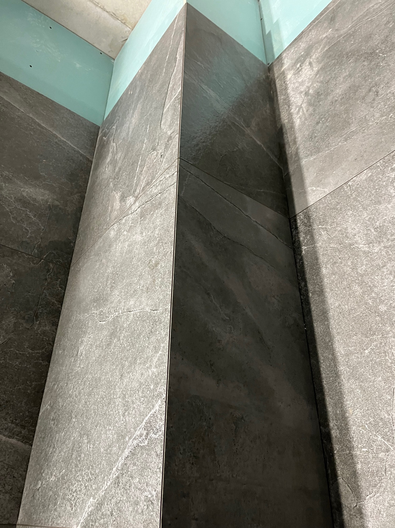 Szare, matowe płytki imitujące kamień, ułożone w rogu łazienki. Widoczne łączenia płytek i fragment sufitu z płyt karton-gips. Ujęcie z dołu, podkreślające pionowość.