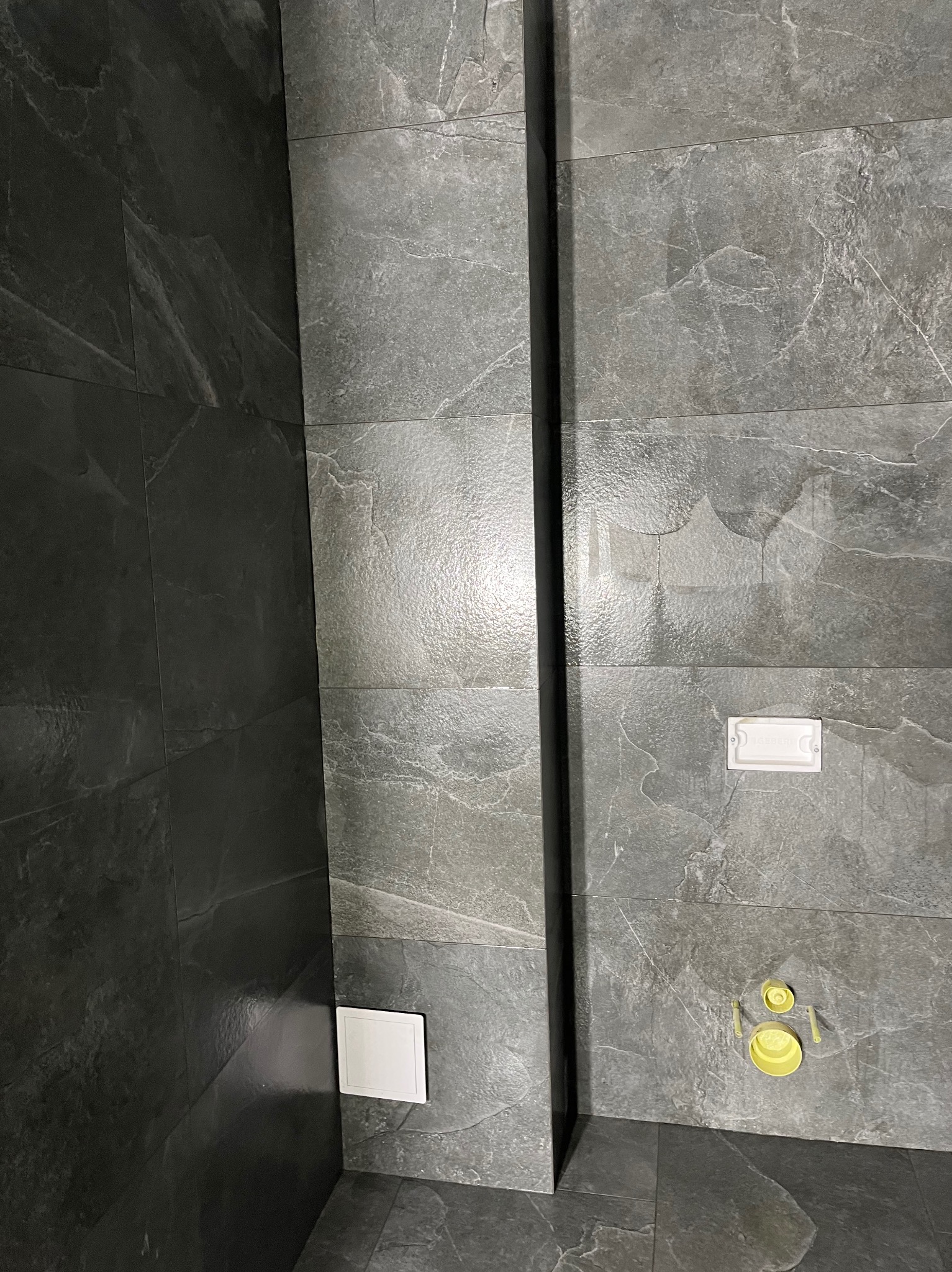 Szare płytki imitujące kamień w łazience, z widocznymi elementami instalacji i wentylacji. Minimalistyczny design, nowoczesne wykończenie, ciemna fuga.