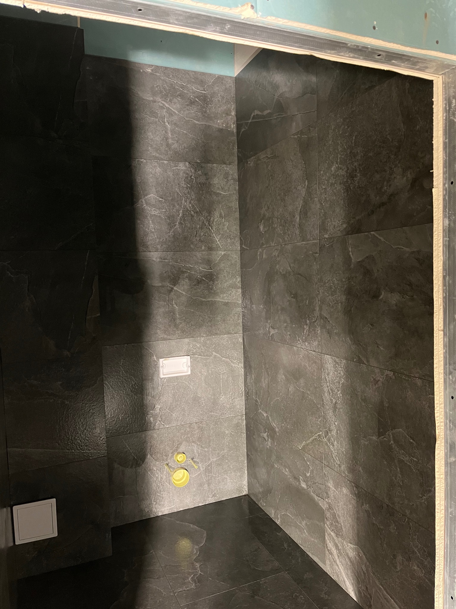 Wykończona łazienka z ciemnymi, marmurkowymi płytkami na ścianach i podłodze. Widoczne elementy instalacji sanitarnej i fragment ościeżnicy.