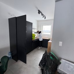 Polsek s.c. PHU. Sklep wielobranżowy. Polehońscy M.T. - Kobieta w białej kurtce sprząta kuchnię z czarnymi szafkami. Na podłodze worki na śmieci i butelka wody. Widok na okno z zimowym krajobrazem.