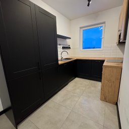 Polsek s.c. PHU. Sklep wielobranżowy. Polehońscy M.T. - Nowoczesna kuchnia w odcieniach czerni i drewna z oknem, minimalistyczne oświetlenie punktowe, jasna podłoga. Widok na fragment pomieszczenia po remoncie.