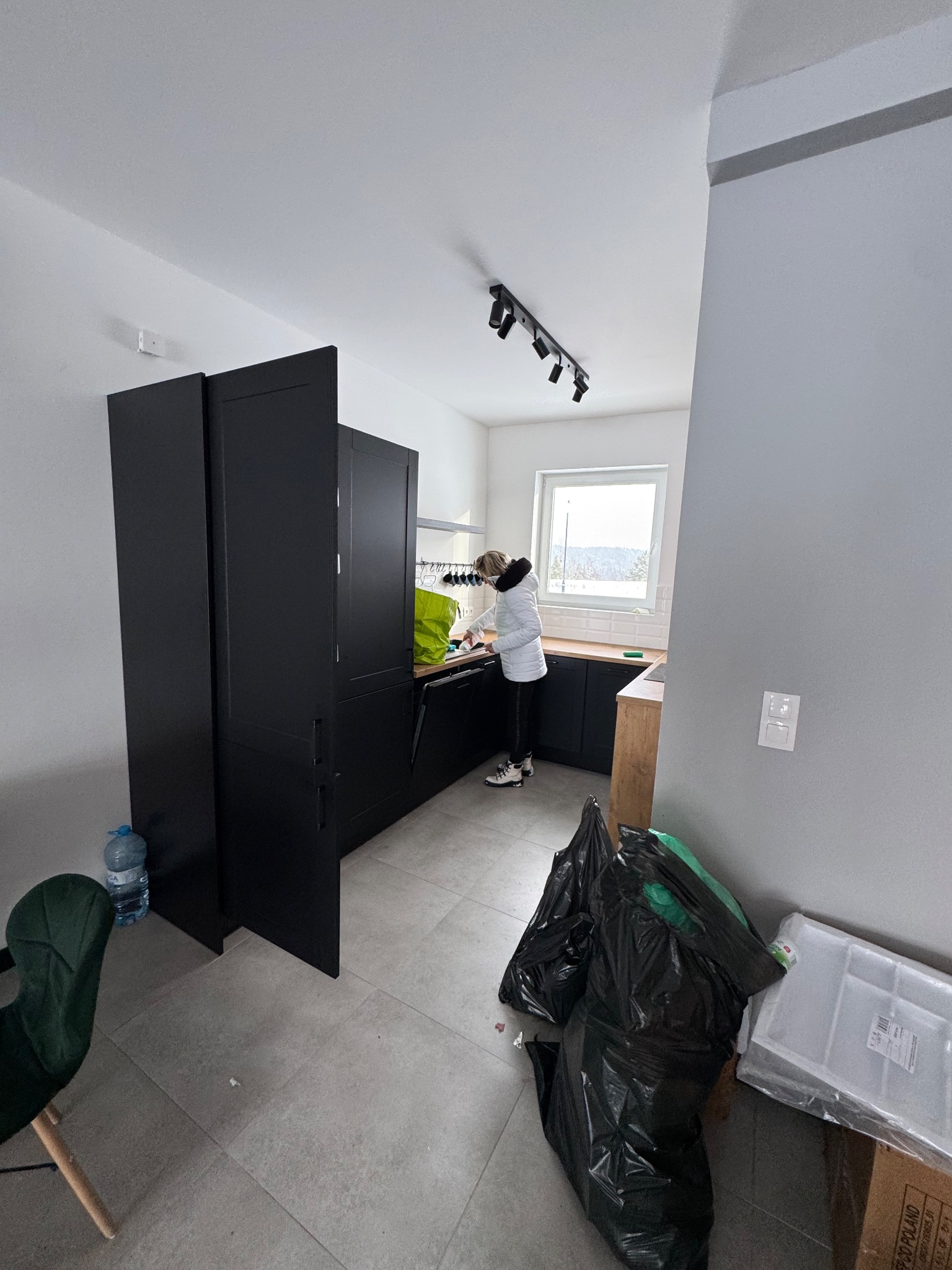 Kobieta w białej kurtce sprząta kuchnię z czarnymi szafkami. Na podłodze worki na śmieci i butelka wody. Widok na okno z zimowym krajobrazem.