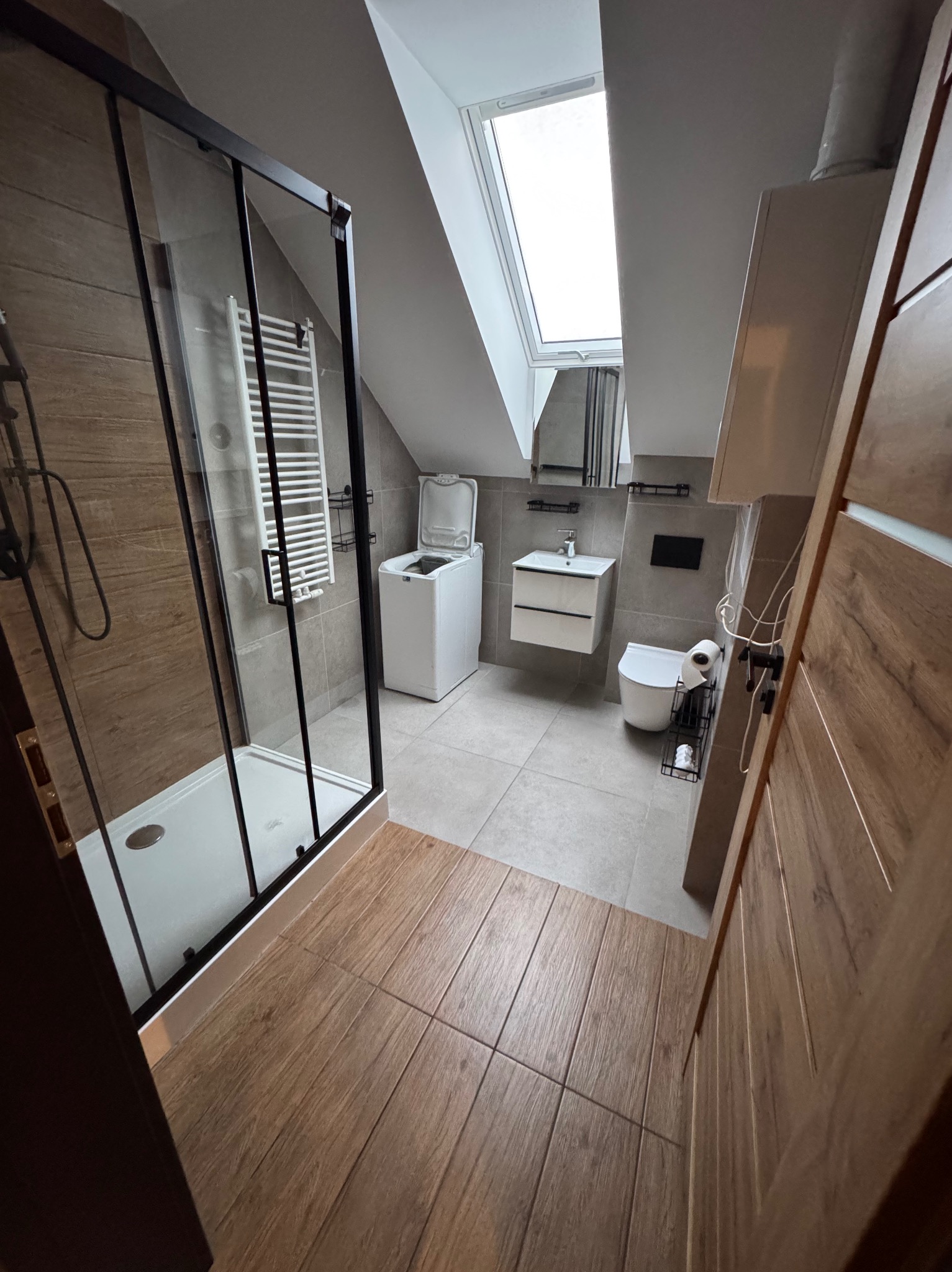 Jasna łazienka na poddaszu z kabiną prysznicową, pralką i oknem dachowym. Uwagę przykuwają płytki imitujące drewno na podłodze i ścianie kabiny.