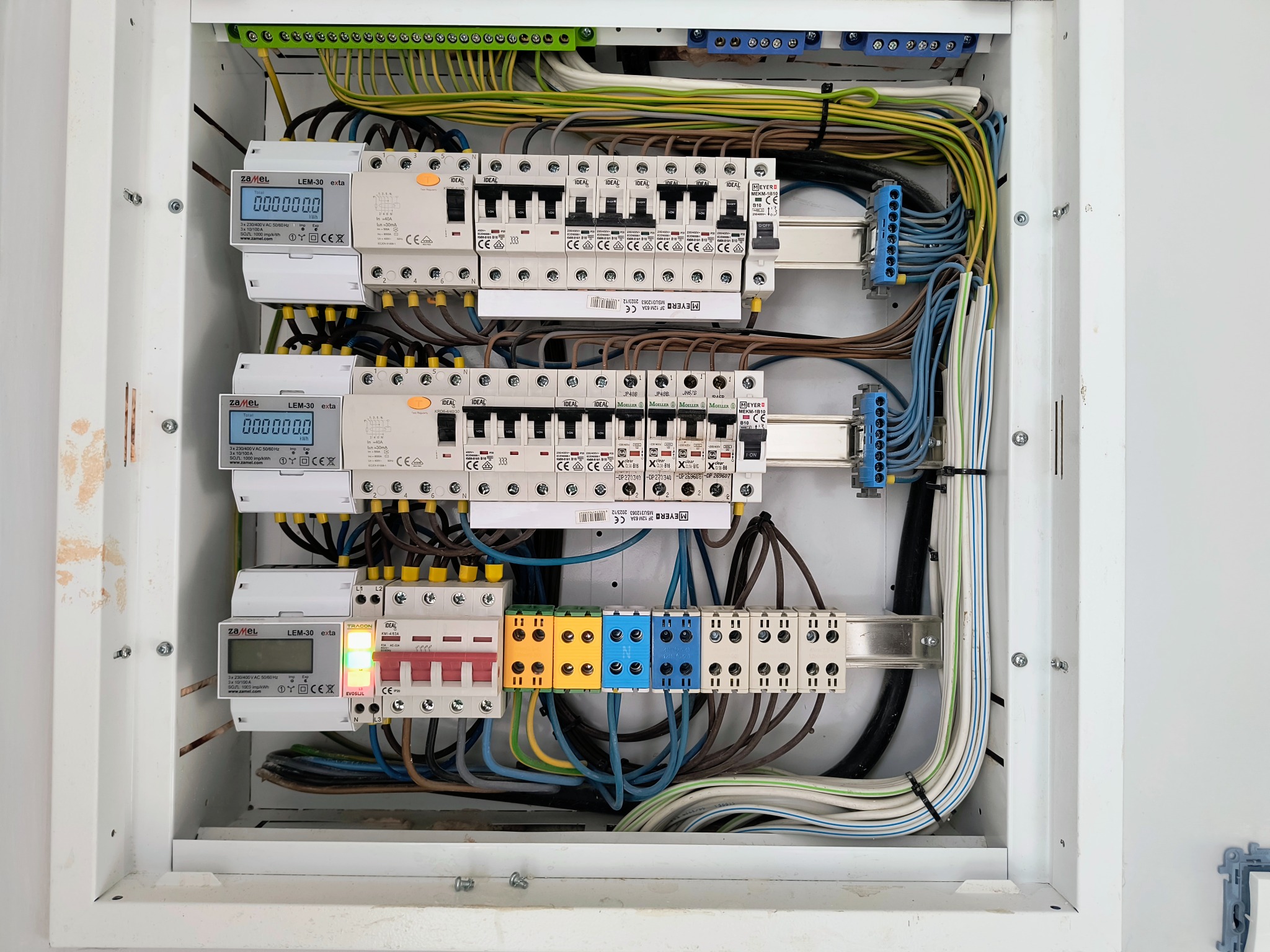 Rozdzielnica elektryczna w białej obudowie, widoczne liczniki, bezpieczniki, przewody w różnych kolorach, starannie ułożone. Instalacja wewnątrz budynku.
