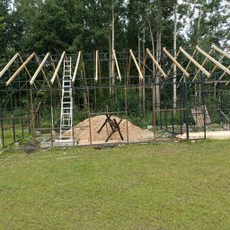 Bartosz N. - Konstrukcja szkieletowa budynku w trakcie budowy, widoczne metalowe ramy, drewniane elementy dachu, drabiny i butla z gazem na trawiastym terenie.