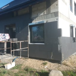 Bartosz N. - Ocieplanie elewacji domu jednorodzinnego czarnym styropianem. Widoczne okna z metalowymi profilami, rusztowanie i materiały budowlane. Etap prac.