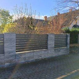 Bartosz N. - Nowoczesne ogrodzenie z szarego kamienia i poziomych, grafitowych paneli, oddzielające posesję od chodnika z kostki brukowej. W tle dom z ciemnym dachem i drzewa.