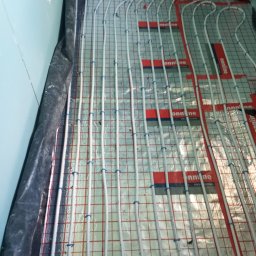 Bartosz N. - Ułożone rury ogrzewania podłogowego na folii izolacyjnej z siatką, zabezpieczone niebieskimi uchwytami, widoczne elementy instalacji na ścianie.