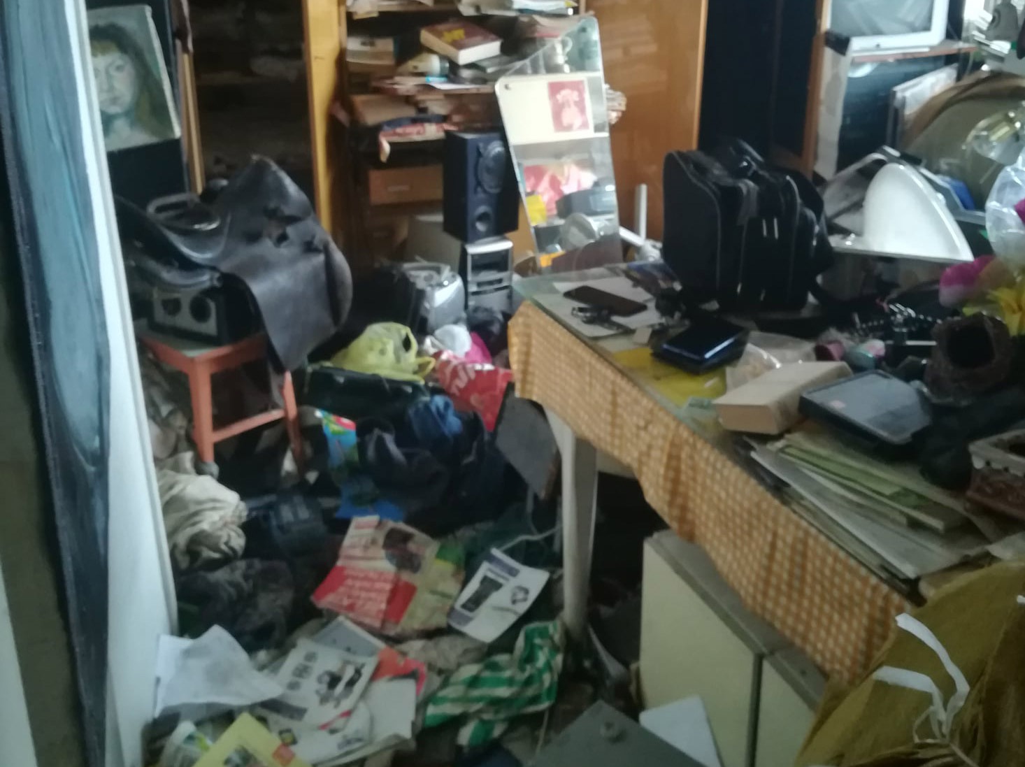 Zabałaganione wnętrze pokoju z różnymi przedmiotami: ubrania, książki, elektronika, gazety, torby, meble. Widoczny obraz na ścianie i stół przykryty obrusem.