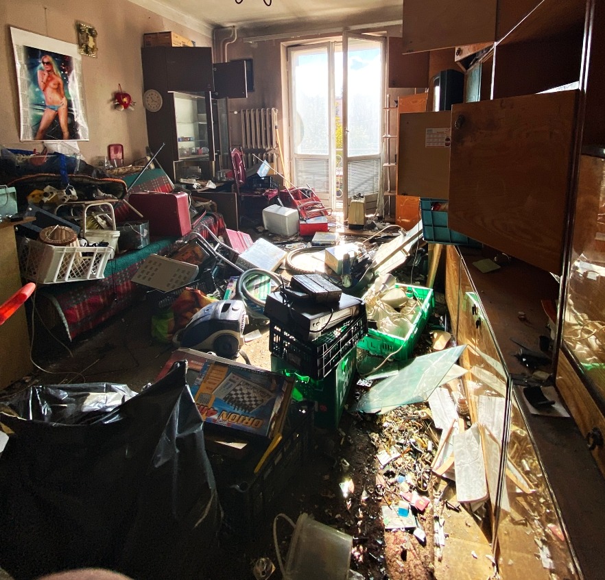 Zrujnowane wnętrze mieszkania z chaotycznym nagromadzeniem przedmiotów, od mebli po sprzęt AGD, w jasnym świetle wpadającym przez balkon. Widoczny bałagan po opróżnianiu.