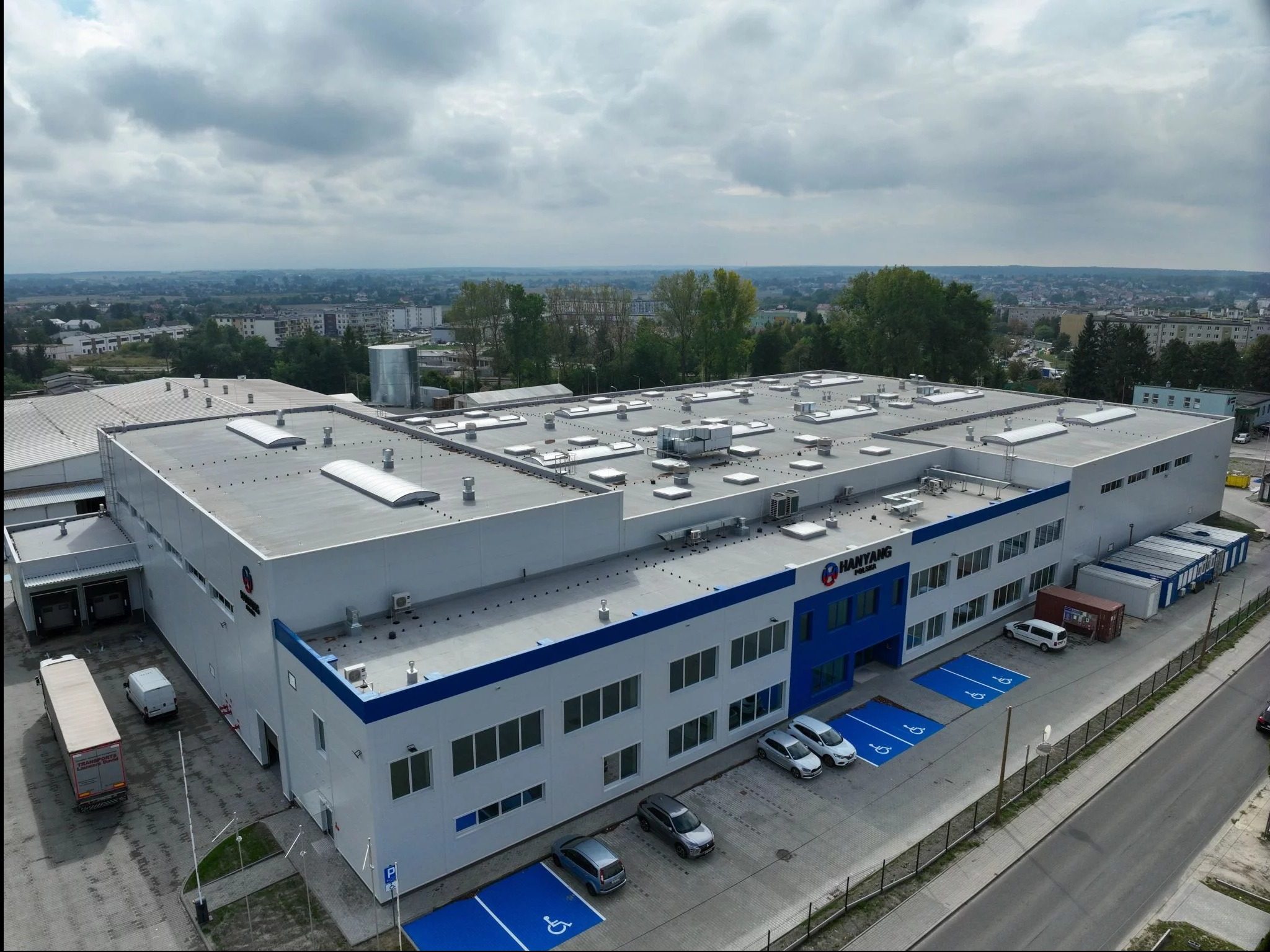 Szeroki kadr z lotu ptaka na nowoczesny budynek przemysłowy z szarym dachem, białymi ścianami i niebieskimi akcentami. Na parkingu zaparkowane samochody i kontenery.