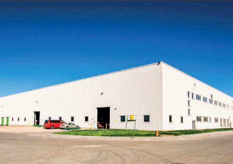 Duży, biały budynek magazynowy z blachy trapezowej pod błękitnym niebem. Widoczne samochody i zieleń. Minimalistyczna architektura przemysłowa.