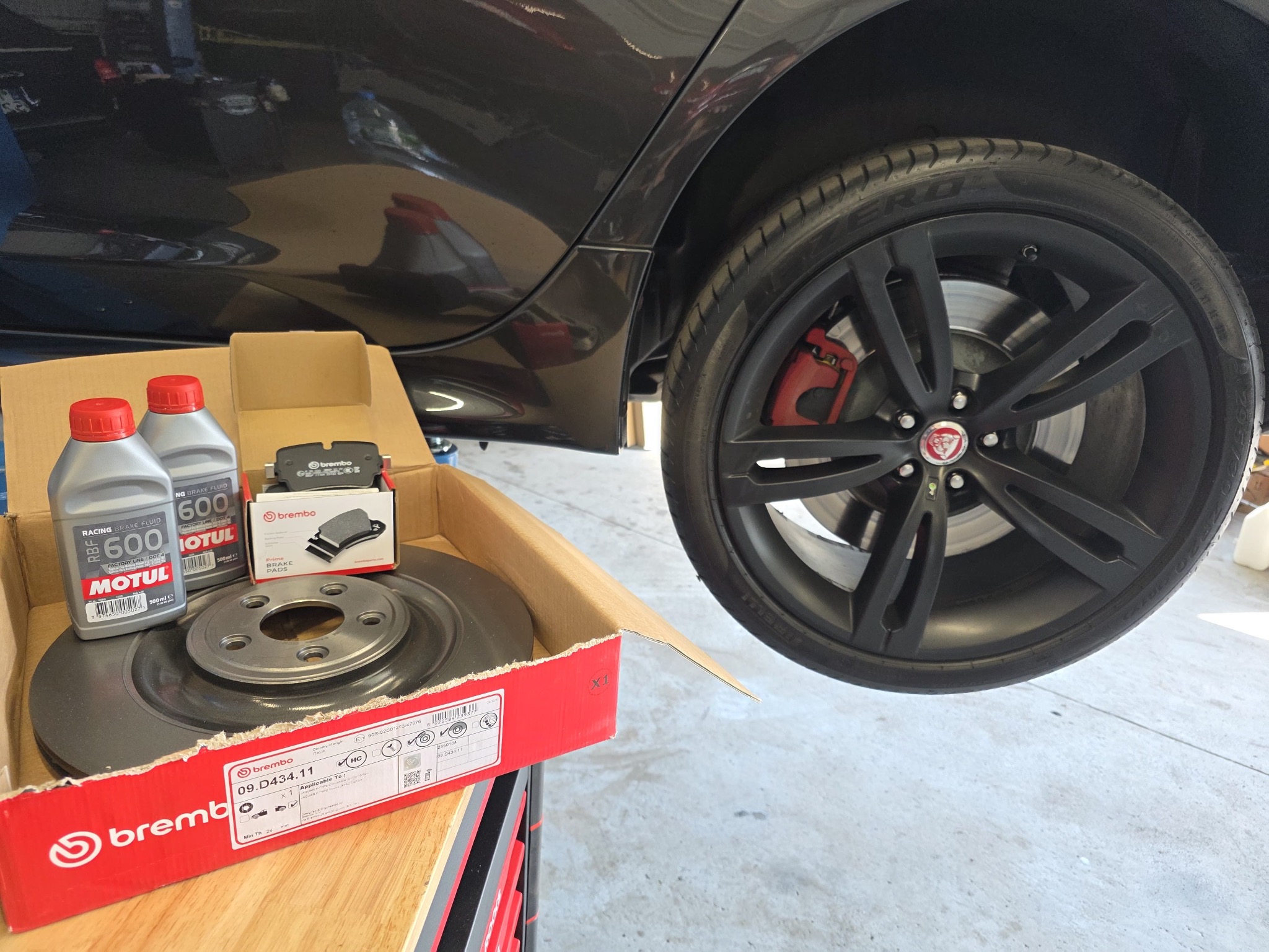 Wymiana tarcz i klocków hamulcowych Brembo w samochodzie Jaguar. Widoczne nowe części, płyn hamulcowy Motul i koło z zaciskiem Brembo. Detal warsztatu.