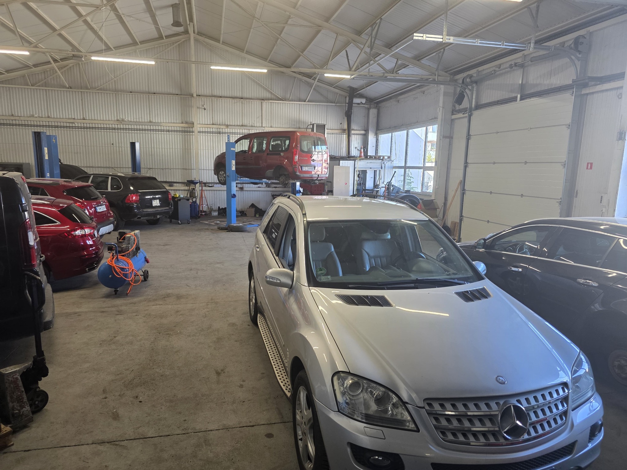 Wnętrze warsztatu samochodowego z podnośnikami i kilkoma autami, w tym srebrny SUV na pierwszym planie. Widoczny kompresor i narzędzia.