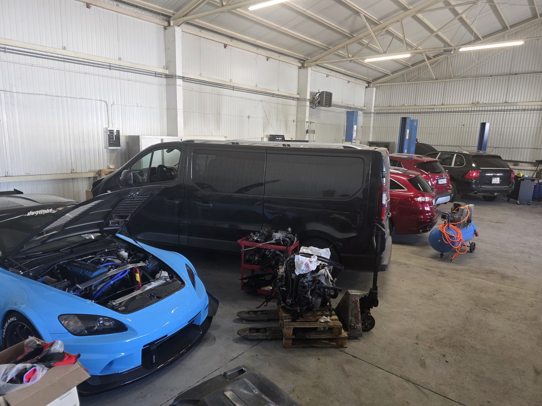 Wnętrze warsztatu: błękitny sportowy samochód z otwartą maską, silnik na palecie, czarny van i czerwone auta w tle. Widoczny kompresor i podnośniki.
