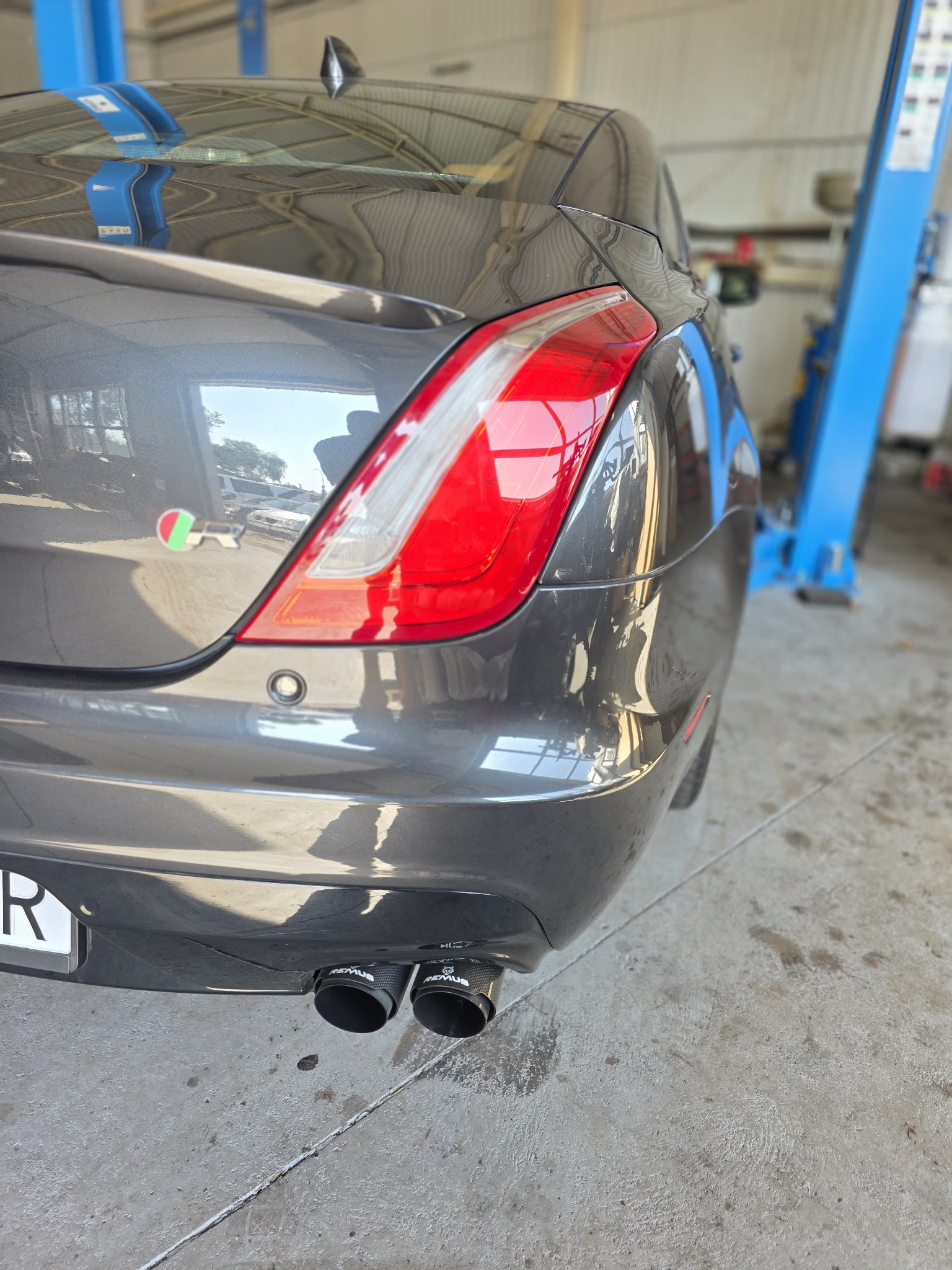Szary Jaguar XJ po tuningu wydechu Remus w warsztacie. Widoczne podwójne końcówki wydechu, tylne światło i podnośnik warsztatowy w tle.