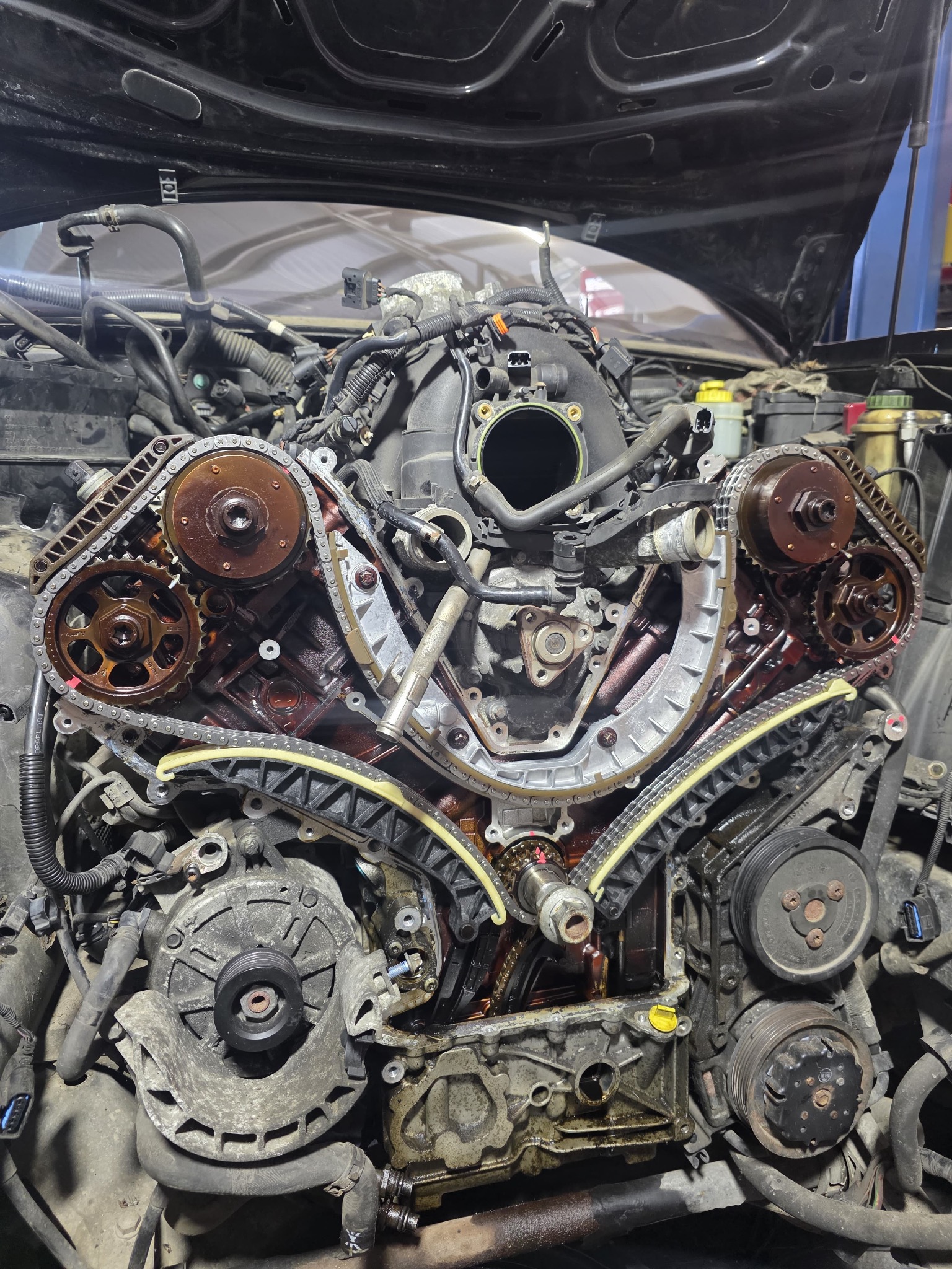 Otwarty silnik samochodowy z widocznym łańcuchem rozrządu i elementami osprzętu, w trakcie naprawy w warsztacie. Widoczne ślady oleju.