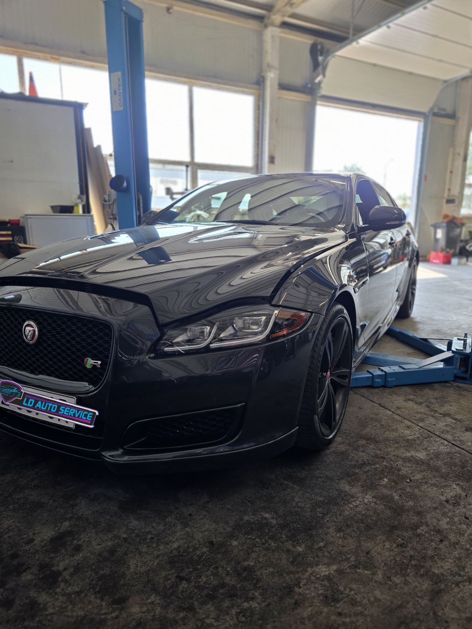 Czarny Jaguar XJ na podnośniku w warsztacie. Widoczne logo serwisu LD Auto Service. Otwarta maska sugeruje trwające prace. Jasne światło wpadające przez bramę.