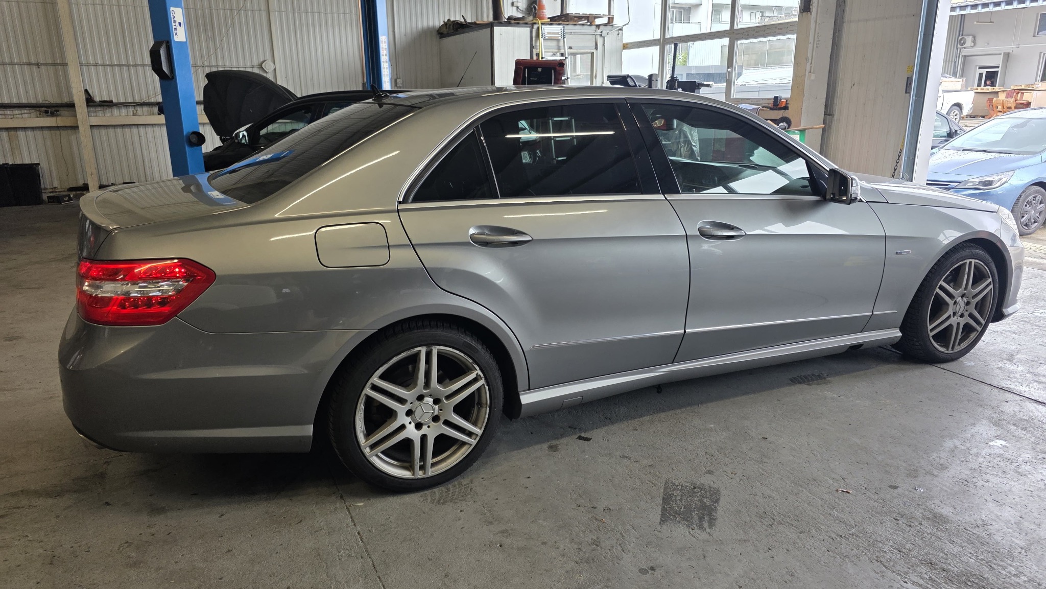 Szary sedan Mercedes-Benz w warsztacie samochodowym, widok z boku, z podniesioną maską innego auta w tle. Widoczne felgi aluminiowe i elementy karoserii.