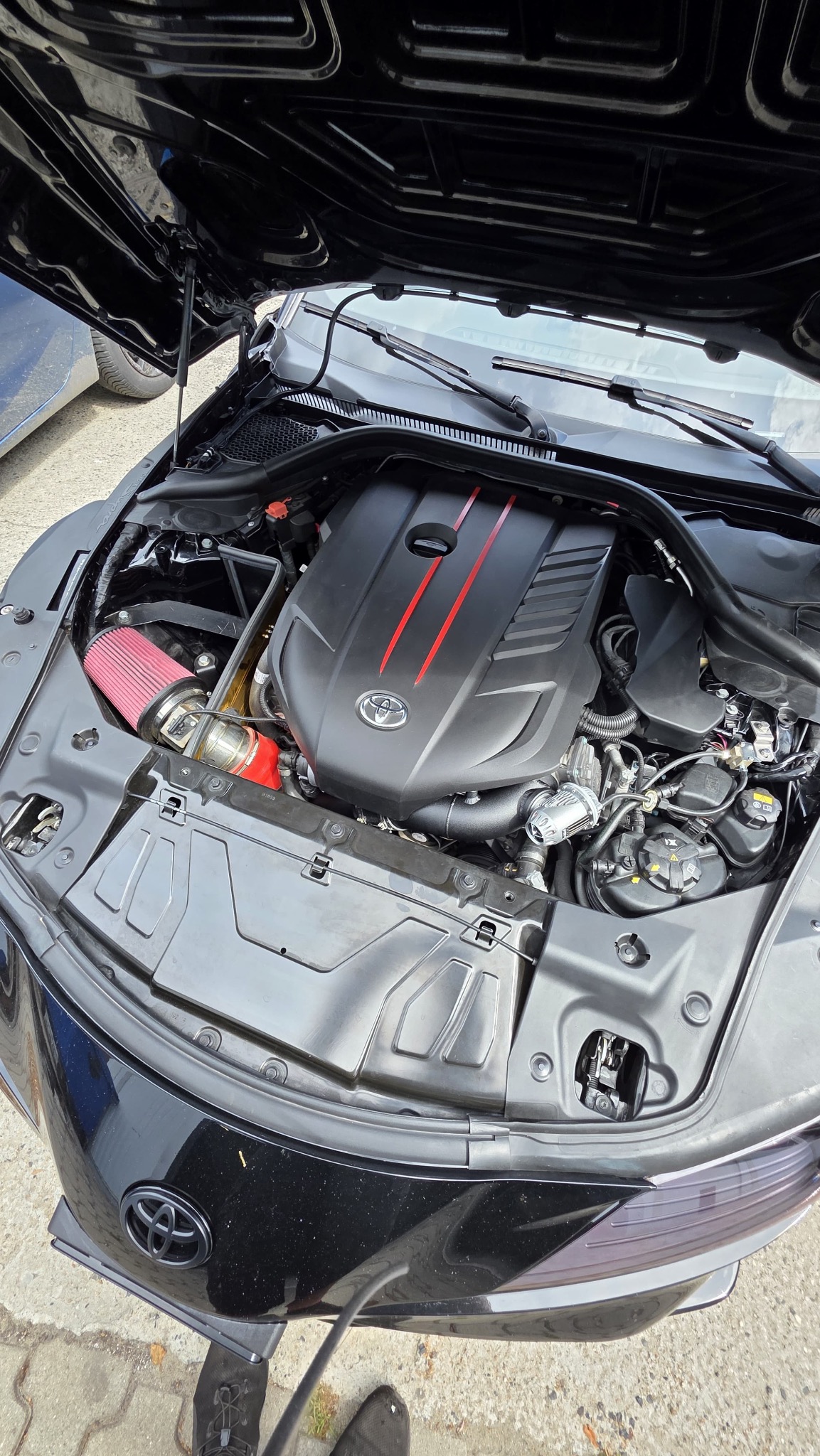 Otwarta maska czarnej Toyoty Supra z widocznym silnikiem z czerwonym filtrem powietrza i elementami w kolorze czerwonym. Widoczne detale silnika i komory.