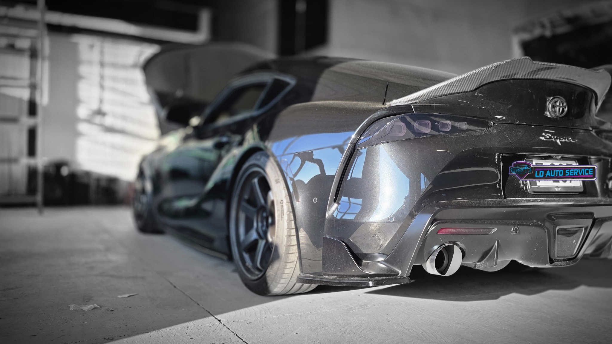 Czarny samochód sportowy Toyota Supra z otwartą maską w warsztacie, widok z tyłu, z logo LD Auto Service na tablicy rejestracyjnej. Widoczne detale karoserii i wydech.