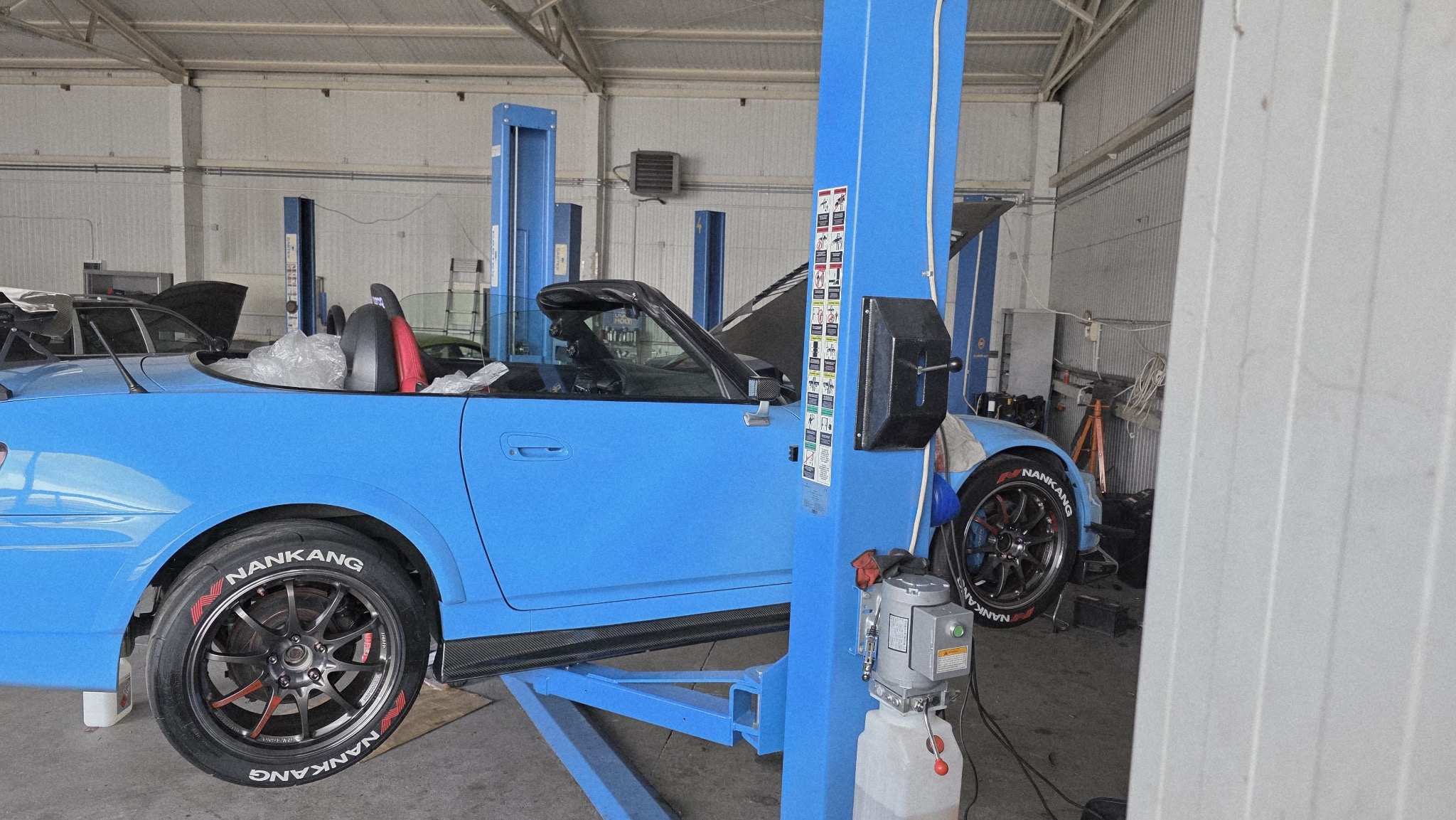 Niebieski kabriolet na podnośniku w warsztacie. Widoczne opony Nankang, podniesiona maska, elementy wyposażenia warsztatu. Jasne oświetlenie, metalowe ściany.