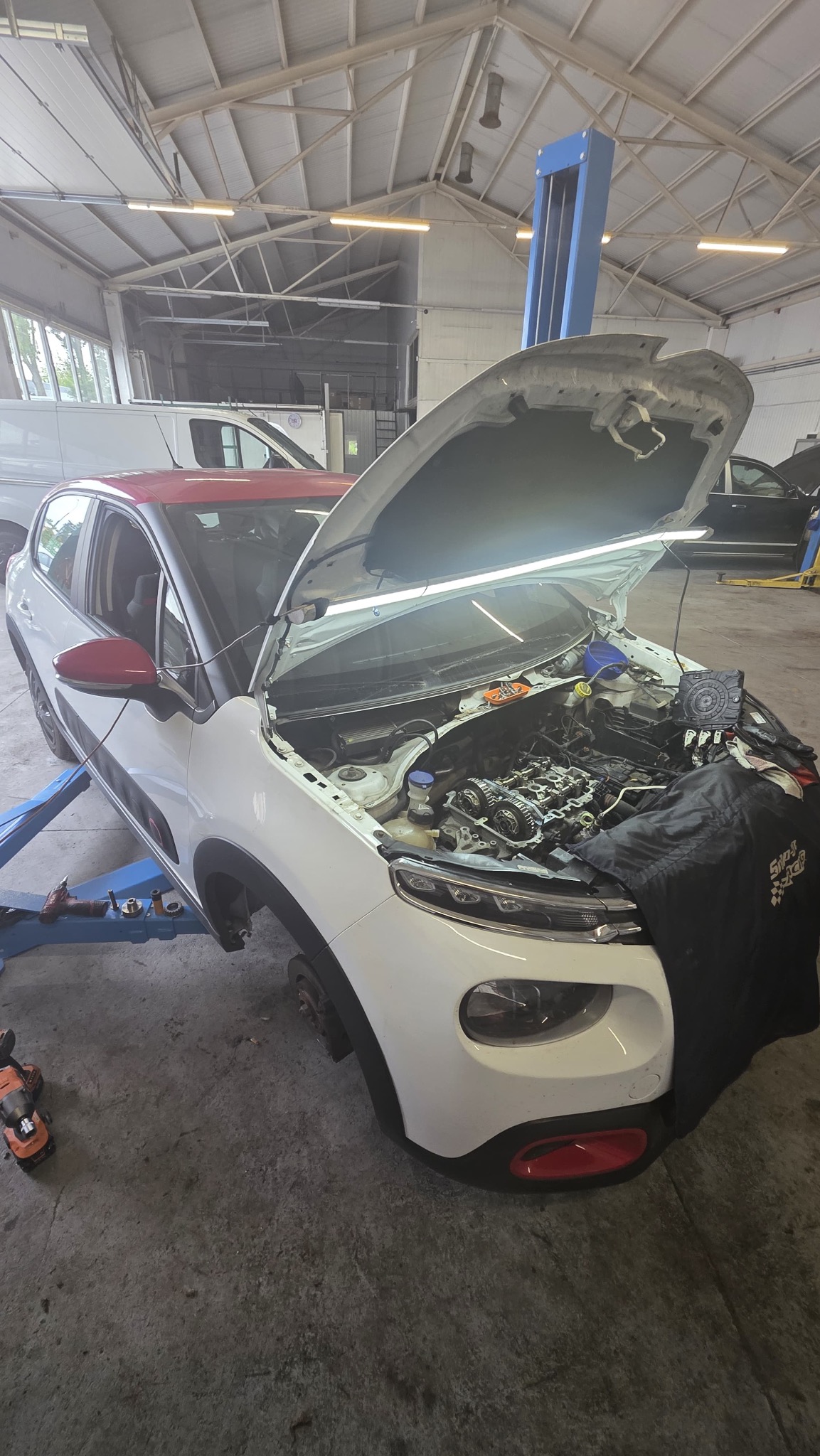 Biało-czerwony samochód na podnośniku w warsztacie z otwartą maską, widoczny silnik i narzędzia. Czarna osłona na błotniku. Brak kół.