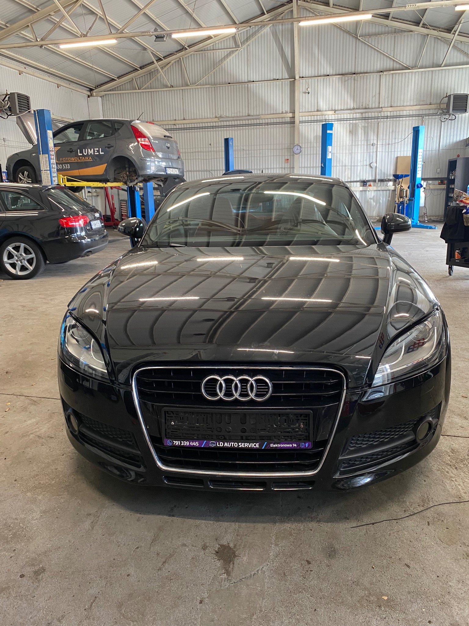 Czarny Audi TT w warsztacie samochodowym, widok z przodu. W tle podnośniki z innymi autami, oświetlenie warsztatowe.