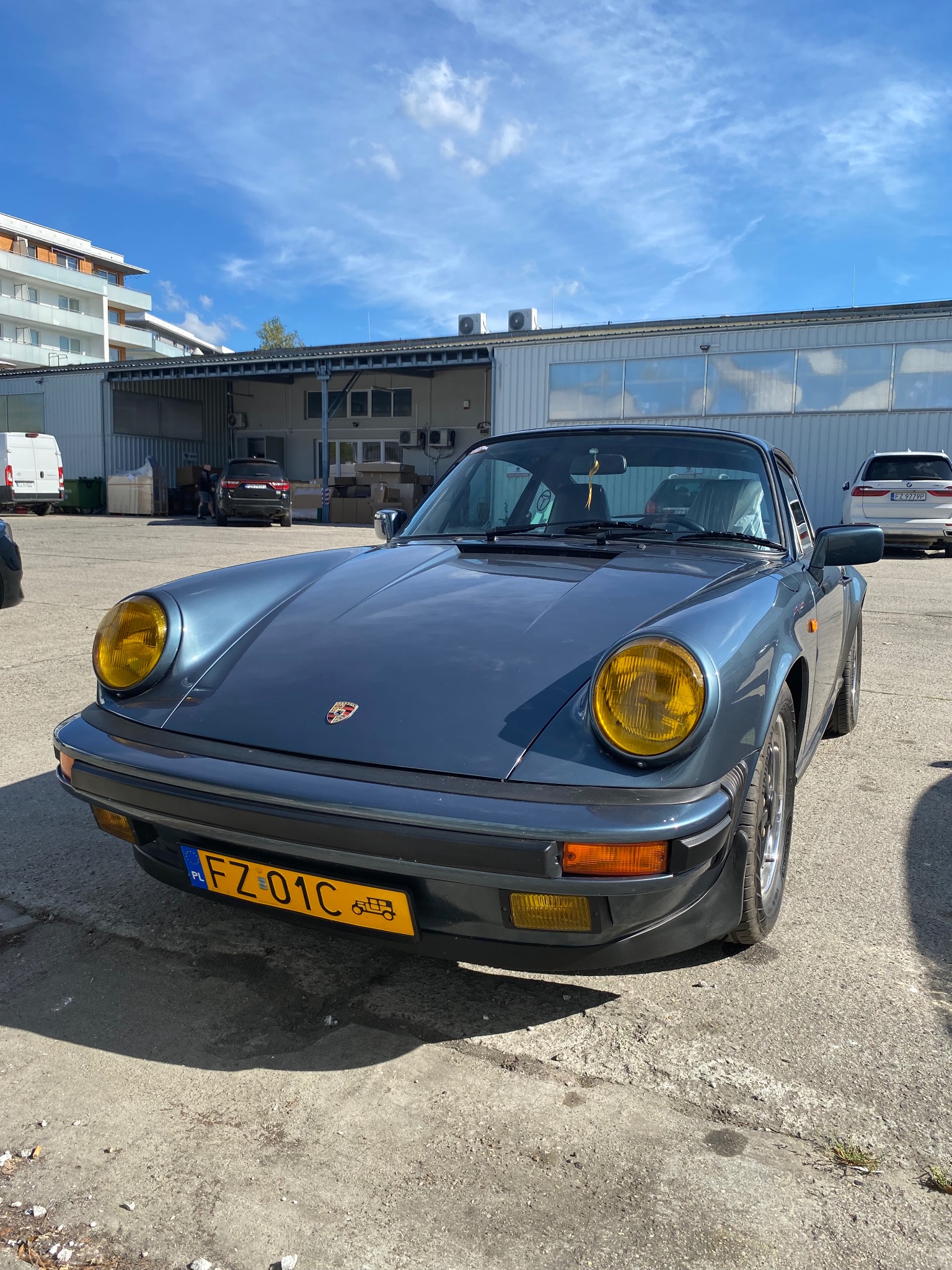 Niebieski, klasyczny Porsche 911 z żółtymi reflektorami na betonowym placu przed warsztatem. Widoczne budynki i inne pojazdy w tle. Tablica rejestracyjna FZ 01C.