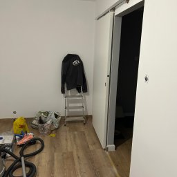 KINS Usługi Remontowe Sławomir Kopek - Wnętrze pokoju w trakcie montażu przesuwnych drzwi. Widoczna drabina, narzędzia i akcesoria malarskie. Podłoga wyłożona panelami imitującymi drewno.