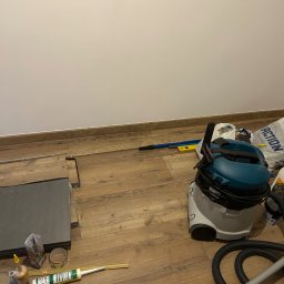KINS Usługi Remontowe Sławomir Kopek - Układanie paneli podłogowych w trakcie remontu, widoczne narzędzia, odkurzacz przemysłowy i materiały budowlane na drewnianej podłodze.