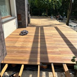 KINS Usługi Remontowe Sławomir Kopek - Nowa drewniana deska tarasowa w trakcie montażu, widoczne legary i podłoże. W tle osoba pracująca przy mieszaniu zaprawy. Słoneczny dzień, widok na ogród.